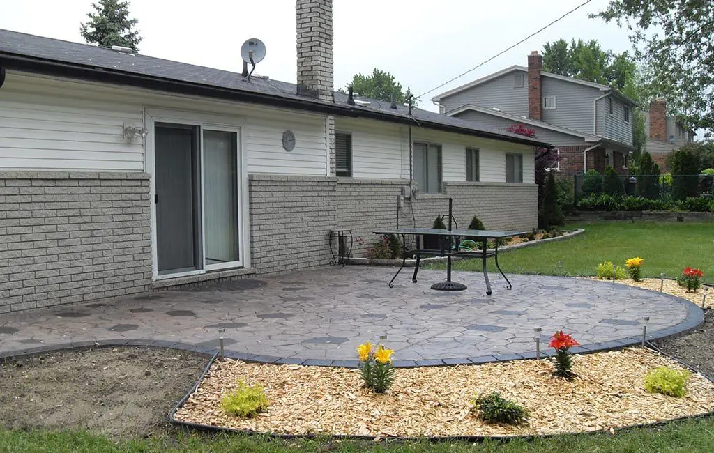 Backyard with a large patterned gray stone patio, a metal table, and a house featuring white siding, gray brick, and a sliding glass door. Mulched garden beds with colorful flowers line the patio edge, with neighboring houses and trees in the background.