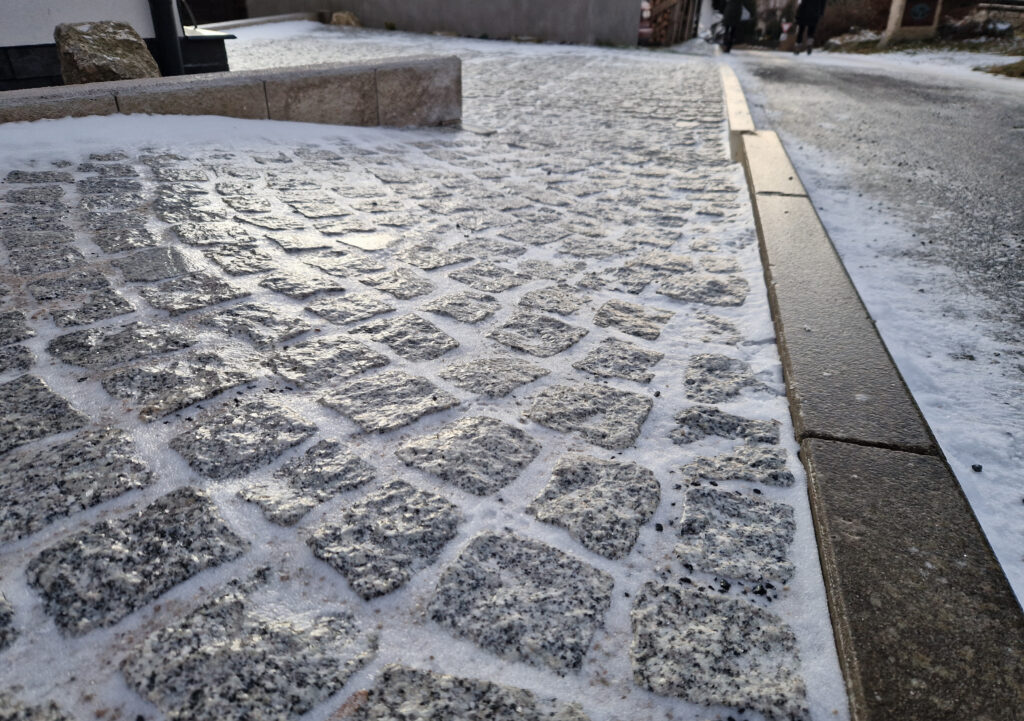 A fine layer of ice causes a shine on a stamped concrete patio; snow is lodged in between the paver joints.