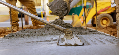 Worker spreading wet concrete with a long-handled float tool while a chute pours more, with construction equipment in the background.