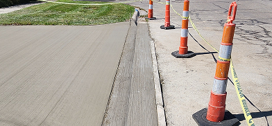 Fresh concrete pour, possibly a driveway or path, with orange construction cones and yellow caution tape.