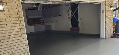 Empty garage with a smooth, light gray floor and light-colored walls. Gray wall-mounted cabinets are on the back wall, and a dark door with yellow tape forming an X is on the right. Brick pillars with outdoor lights flank the open garage entrance.