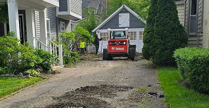 Bobcat loader and workers demolishing a cracked asphalt driveway in front of a gray garage.
