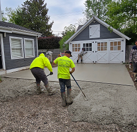 Construction workers in neon shirts pouring and leveling concrete for a new driveway next to a house and garage.