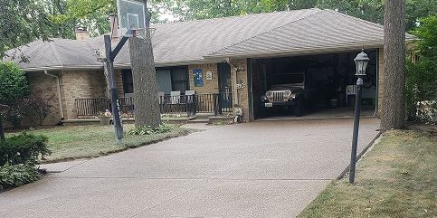 Front view of a brick house with a paved driveway, open garage showing a Jeep, and a basketball hoop in the yard.