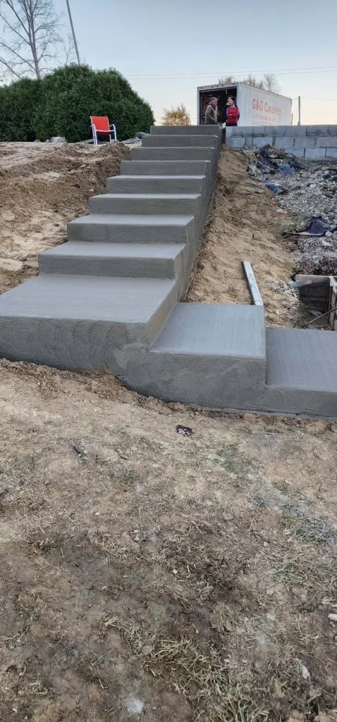 Newly poured concrete steps with a small landing ascend a dirt slope. G&G Concrete truck & workers in background.