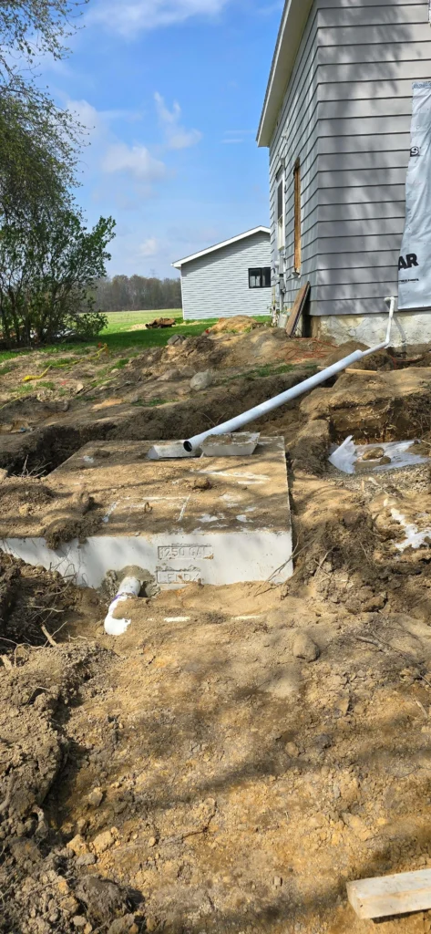 New 1250-gallon septic tank being installed next to a gray-sided house under construction, with connecting pipes in excavated dirt.