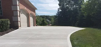 Light-colored concrete driveway leading to a brick house with two arched garage doors, bordered by green grass and trees in the background under a cloudy sky.