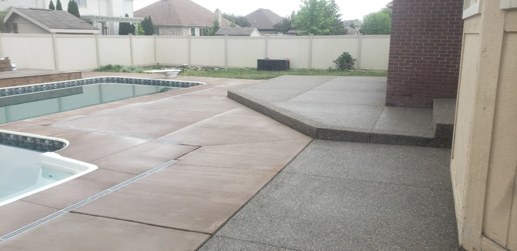 Backyard pool area with light concrete decking, transitioning to a raised, textured gray patio next to a brick wall. Fence beyond.