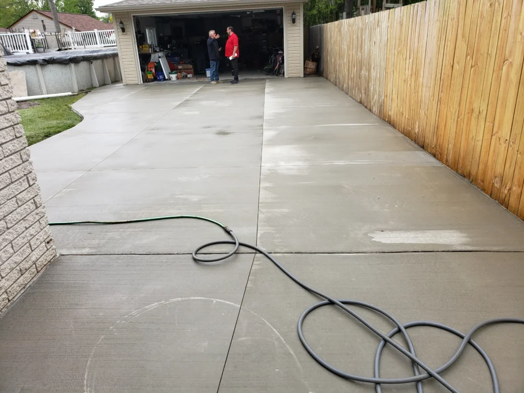 Newly installed concrete driveway and patio, wet. Two men talk by a garage. Features an above-ground pool, wooden fence, and garden hose.