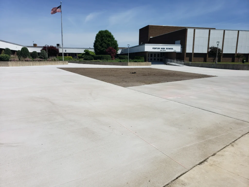 Fenton High School main entry with an American flag, a newly paved concrete plaza, and an unlandscaped dirt area.