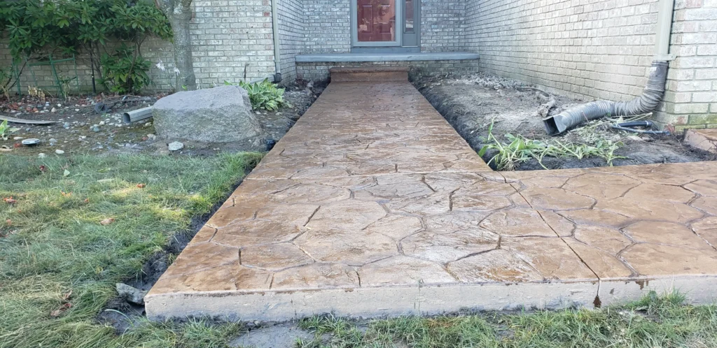 Wet, newly installed stamped concrete walkway with a flagstone pattern leading to a brick house entrance.