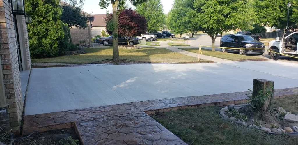 New concrete driveway and stamped walkway by a house. Residential street with parked cars, green trees, and caution tape.