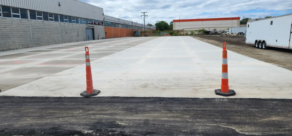Newly paved concrete lot next to fresh asphalt, with two orange traffic cones. Industrial buildings & trailers behind.
