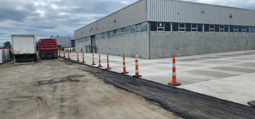 Industrial building, trucks, fresh asphalt patch, and orange cones dividing a dirt path from a concrete loading area.