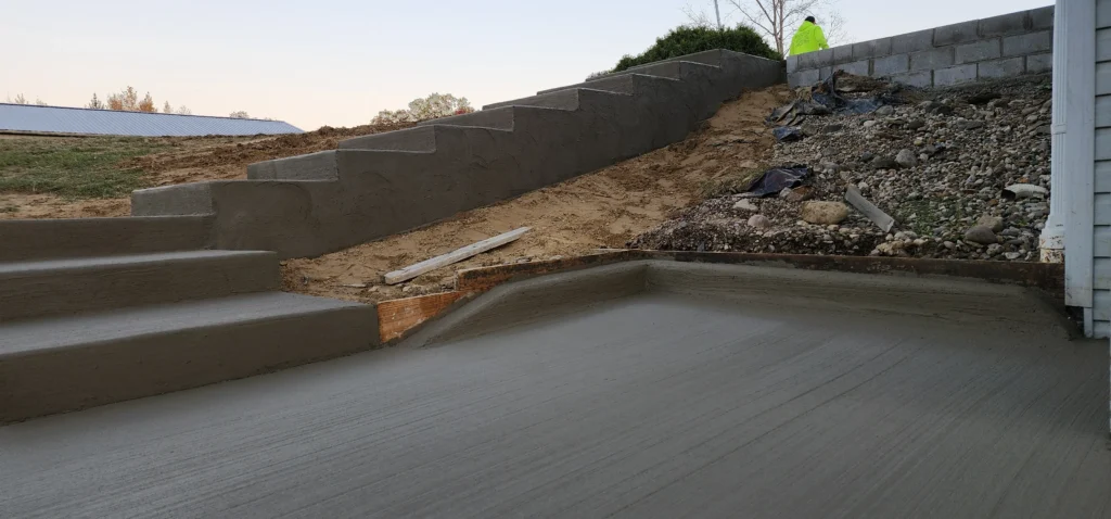 Newly cast concrete steps ascend a dirt embankment to a block retaining wall; a fresh concrete patio is in the foreground.
