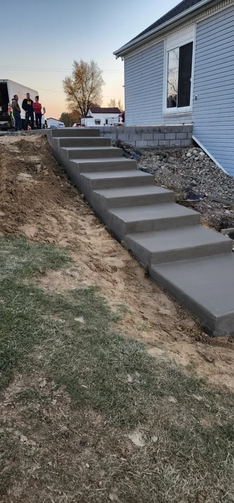 Freshly poured concrete steps built into a dirt slope beside a light blue house. Construction workers stand nearby.