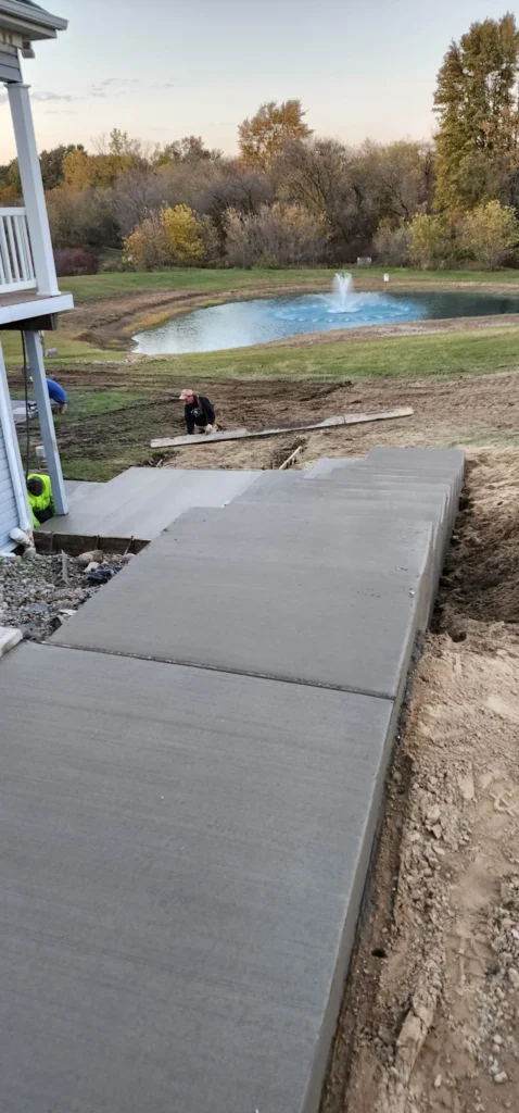 Workers pour concrete for new steps and walkway, overlooking a pond with a fountain and autumn trees.