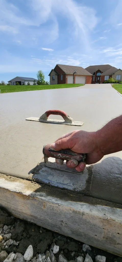 Smoothing a new concrete driveway with a trowel. Worker's hand, another trowel, and homes in a sunny suburban setting.