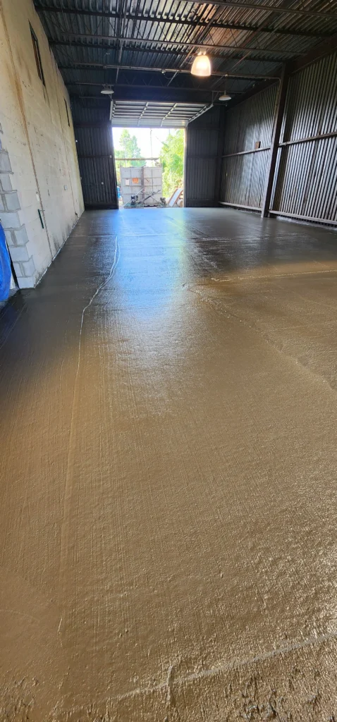 Freshly poured, wet tan concrete floor in an empty warehouse, light streaming from the large open bay door.