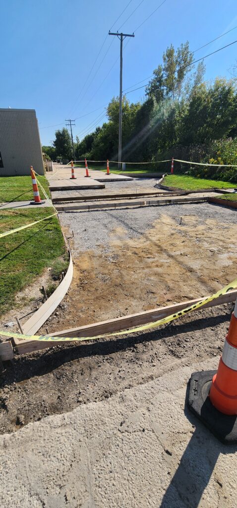 Construction site with wooden forms set for concrete pour, gravel base, safety cones, and yellow caution tape.