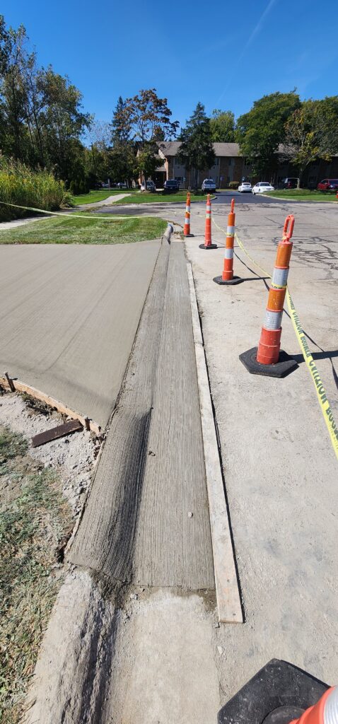 Freshly poured concrete for a new sidewalk or driveway, secured by orange cones and caution tape in a residential neighborhood.