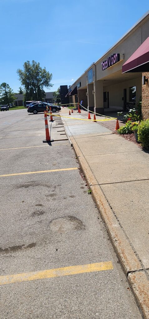 Sidewalk ramp blocked by orange cones and yellow caution tape in front of a strip mall with a 'Total Vision' store.