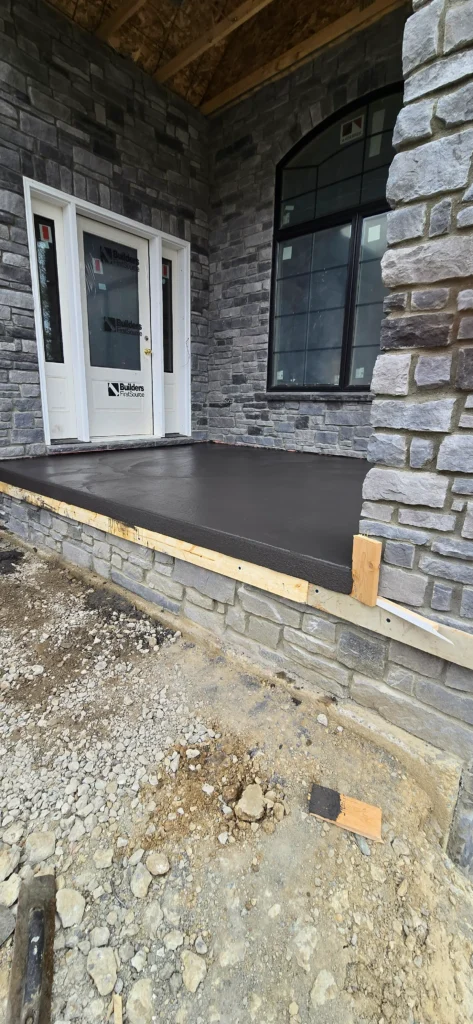 Newly poured dark gray concrete porch and stone facade of a house under construction.