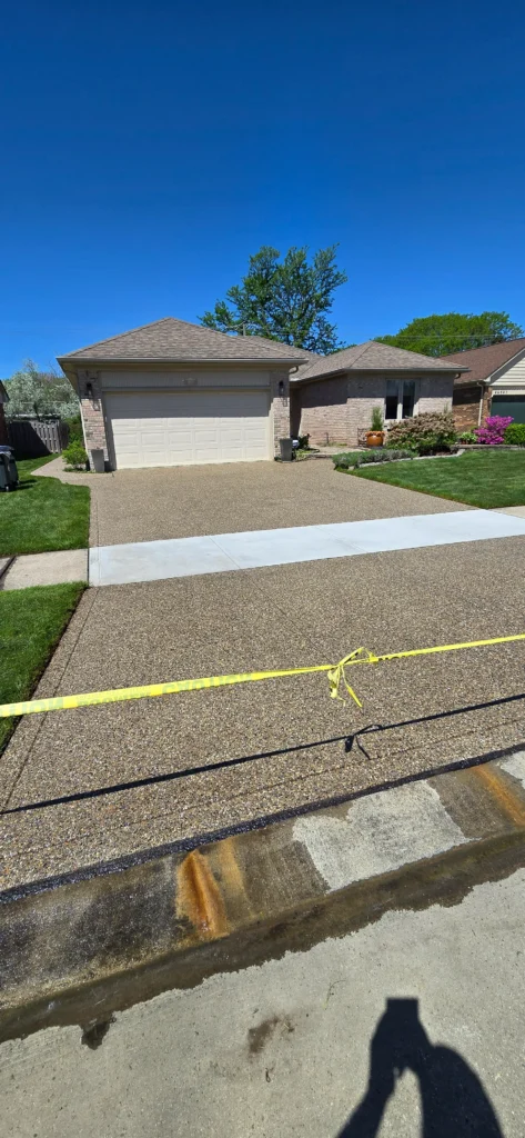 Newly laid exposed aggregate driveway and concrete sidewalk to a brick house, secured by yellow caution tape on a sunny day.