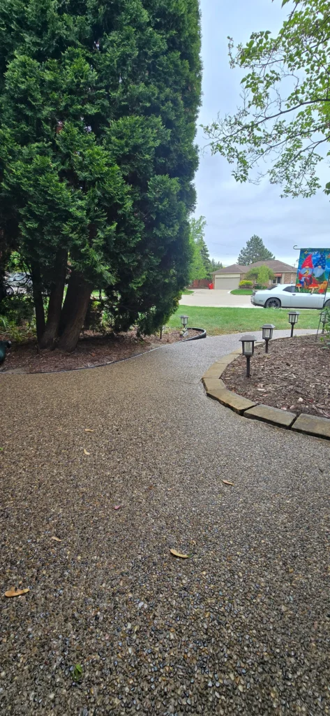 Winding pebble path in a yard with a large evergreen tree, solar lights, and a suburban house with a white car in the distance.