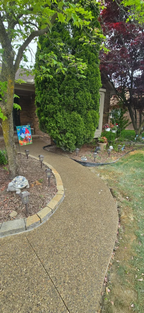 Curved aggregate pathway leading to a brick house, bordered by a garden with a gnome flag, solar lights, and trees.