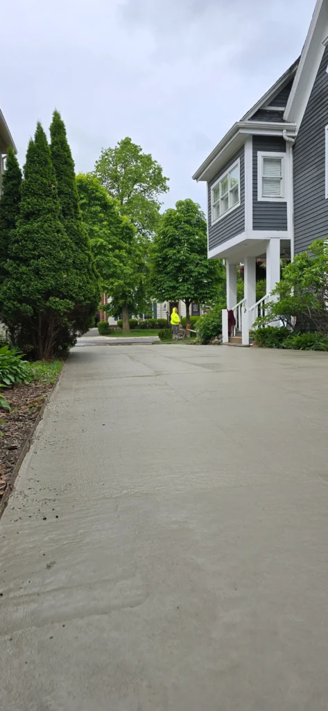 New concrete driveway leading to a gray house, with a worker in yellow and green trees in the background.