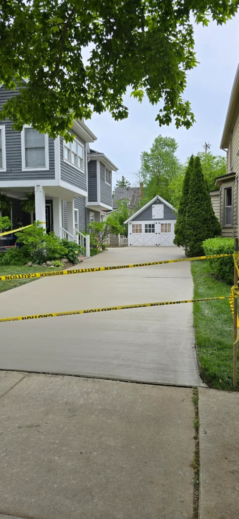 Newly paved concrete driveway with yellow caution tape in front of a gray house and detached garage, under an overcast sky.