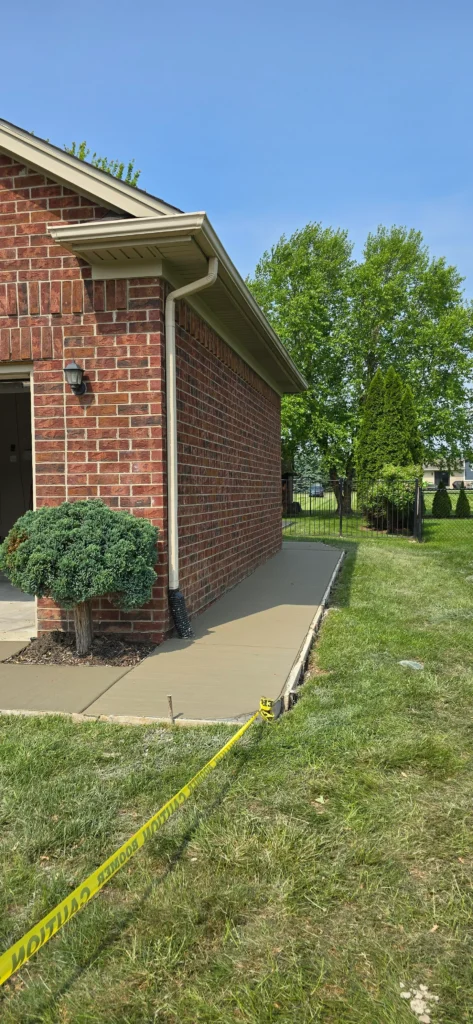 Fresh concrete sidewalk along a brick building, with yellow caution tape across the green grass. Trees and blue sky visible.