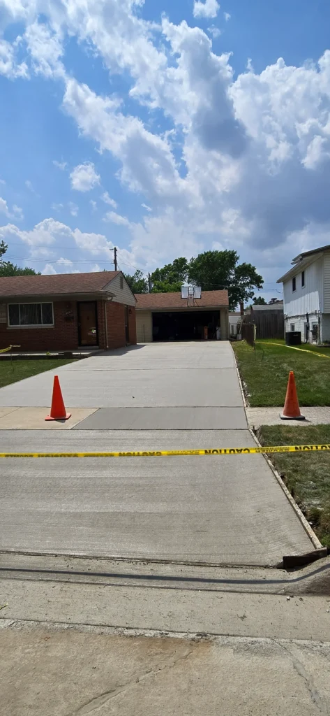 New concrete driveway with caution tape and orange cones leading to a suburban house and garage with a basketball hoop.