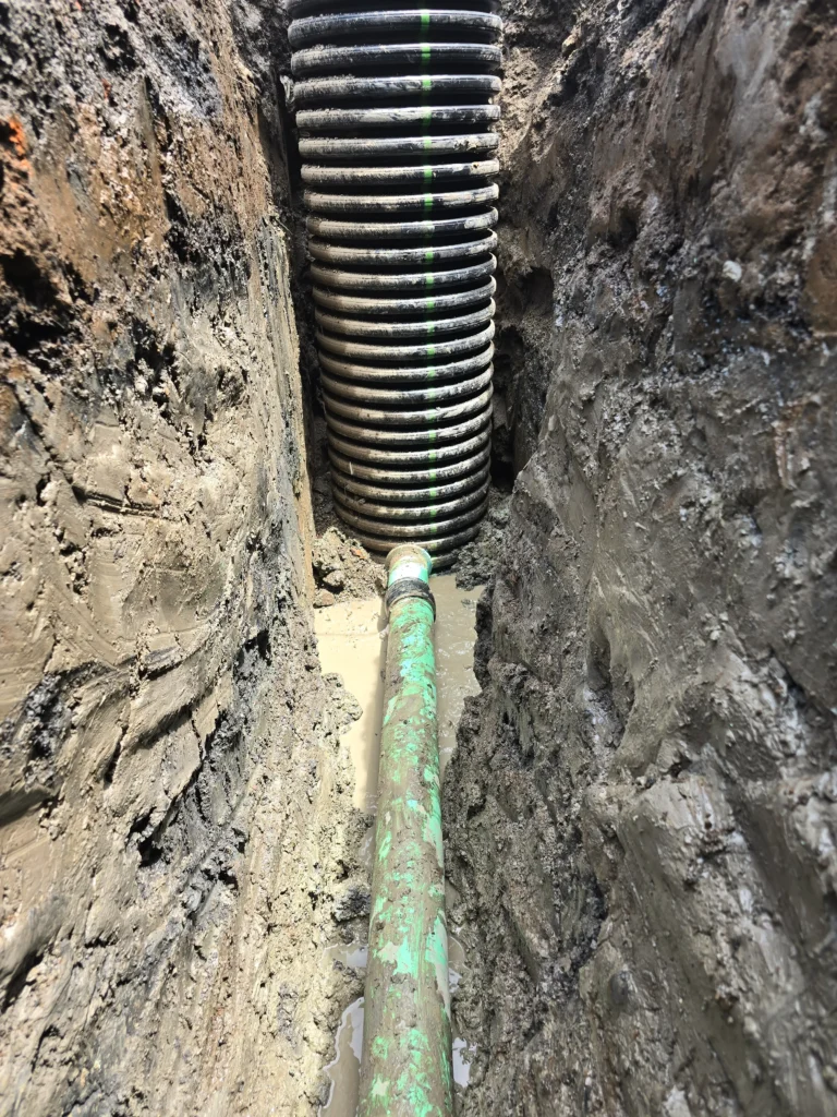 Vertical black corrugated pipe connected to a horizontal green pipe in a muddy excavation trench with standing water.