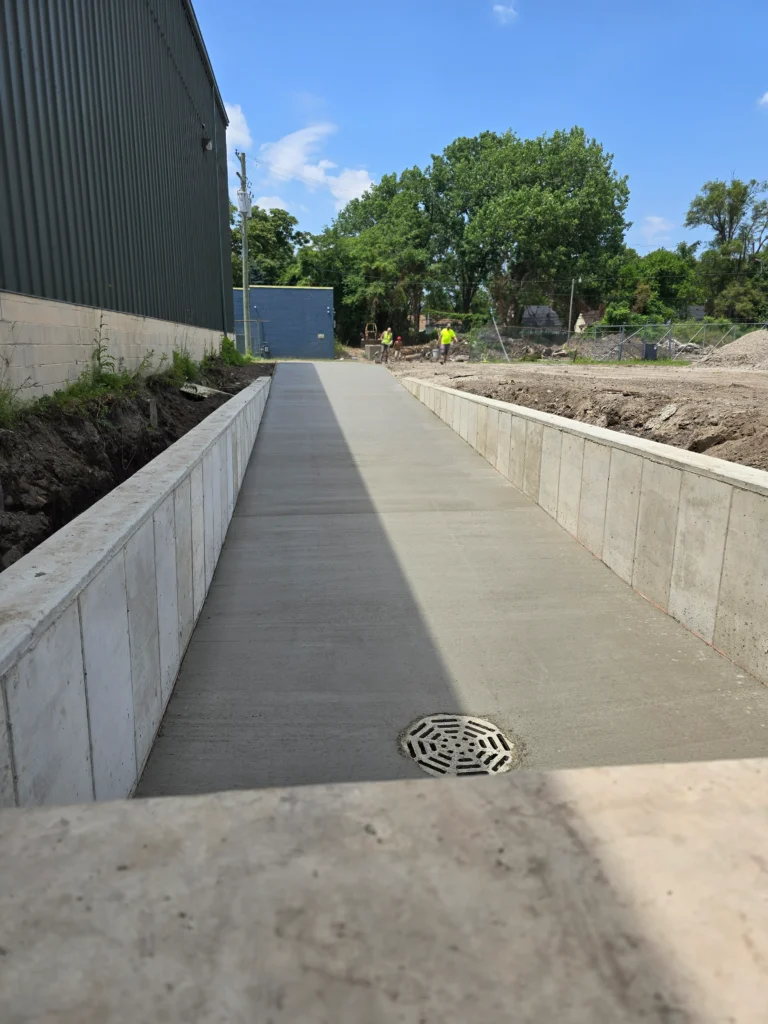 New concrete pathway with retaining walls. On left, a corrugated building; ahead, a construction site with workers. Drain in foreground.