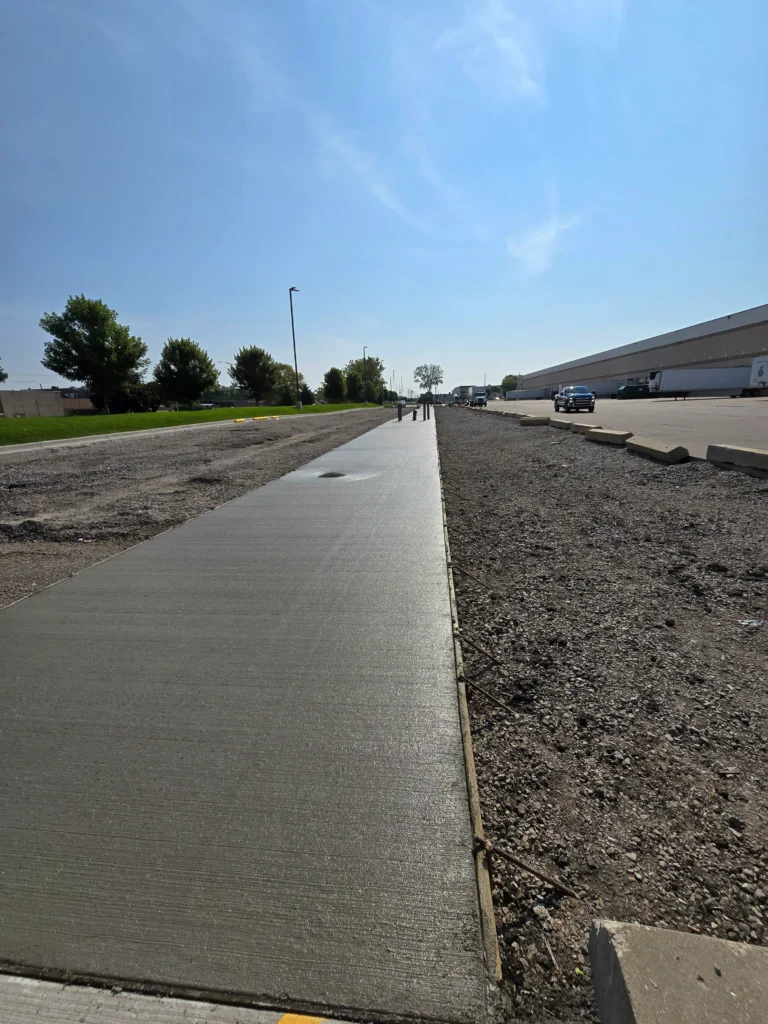 Newly laid concrete sidewalk extends past gravel, trees, and industrial buildings under a clear blue sky.