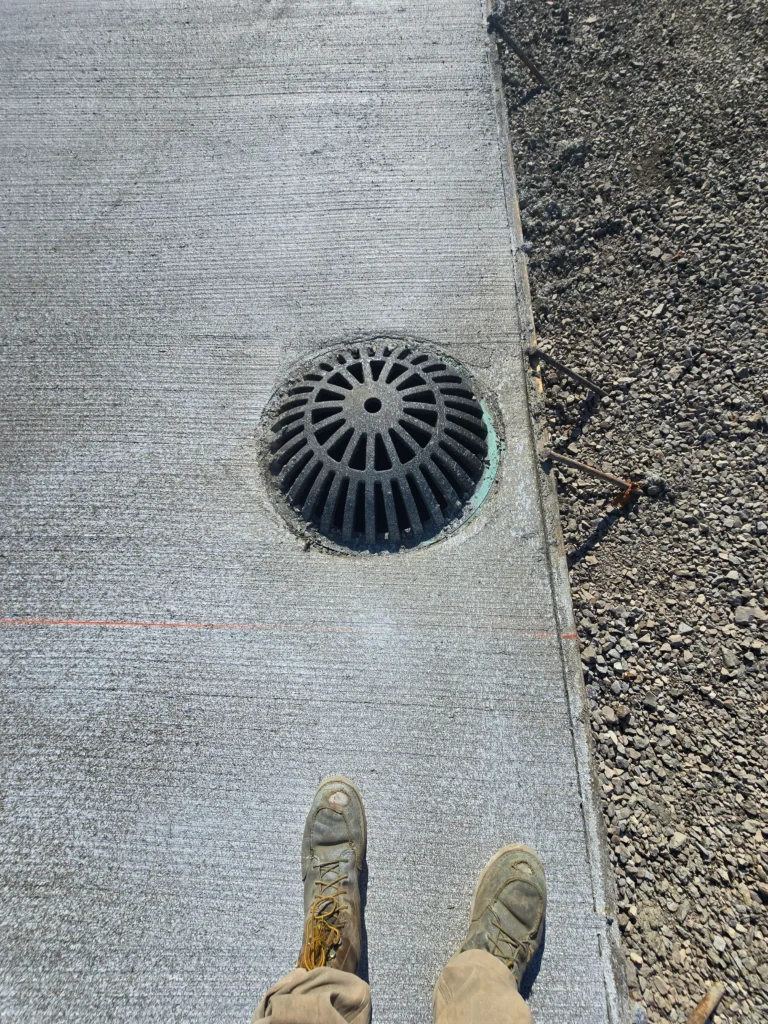 Work boots stand on newly poured, textured concrete with a central drain, a red string line, and a gravel border.