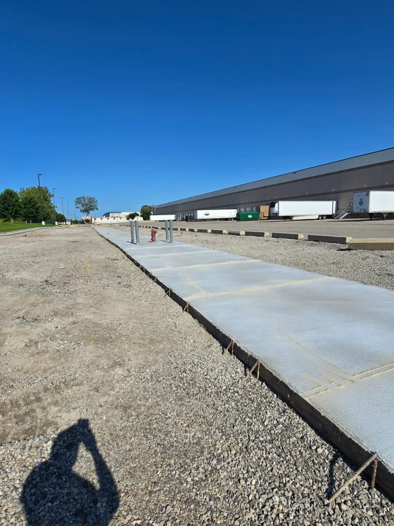 Newly poured concrete sidewalk next to a large warehouse with loading docks, semi-trailers, and a clear blue sky.