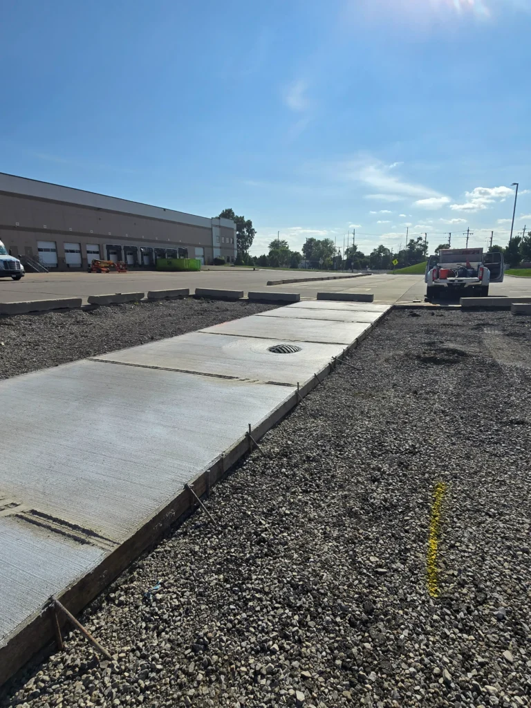 Freshly poured concrete path with a drain, gravel shoulders, leading to a warehouse with loading docks.
