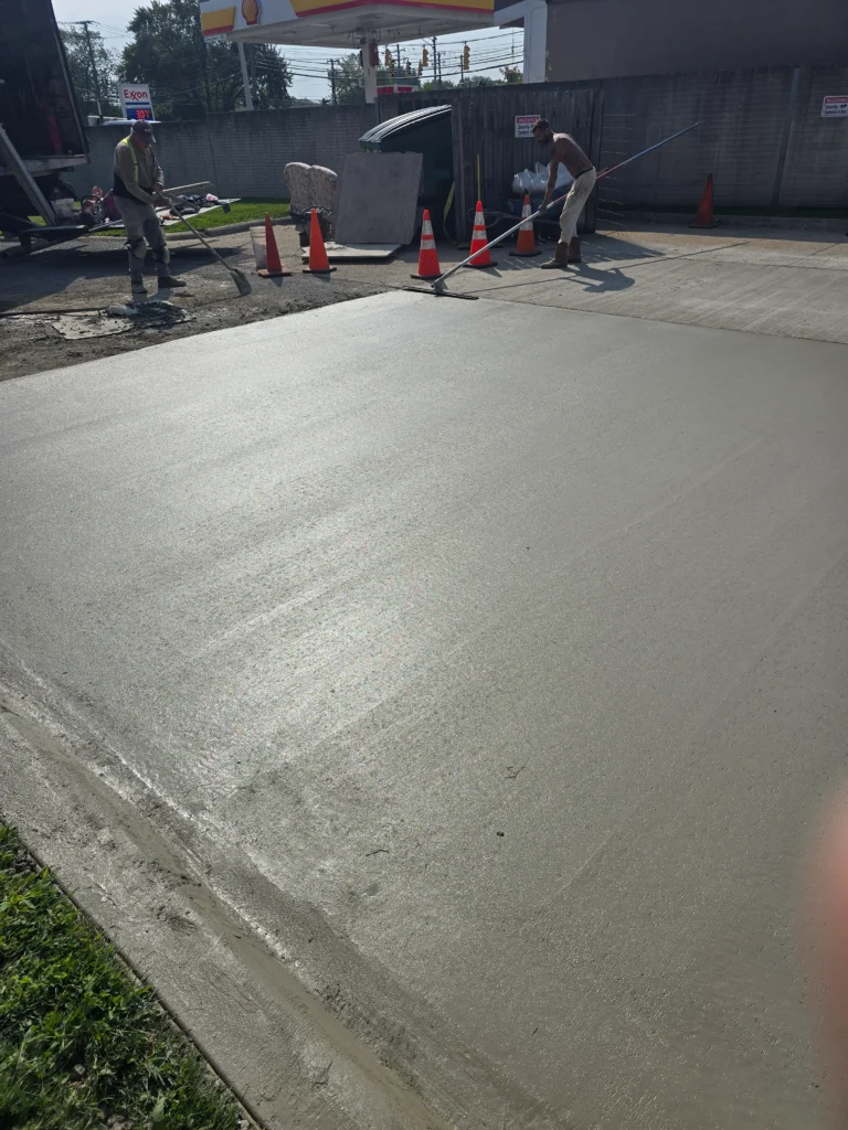 Two workers applying and leveling fresh concrete for a new pavement, with safety cones and a gas station in the background.