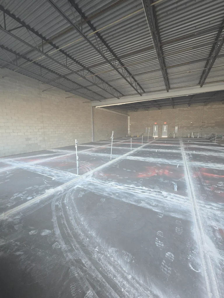 Unfinished commercial space with concrete block walls, exposed ceiling, floor showing grid lines, footprints, and utility stub-outs.