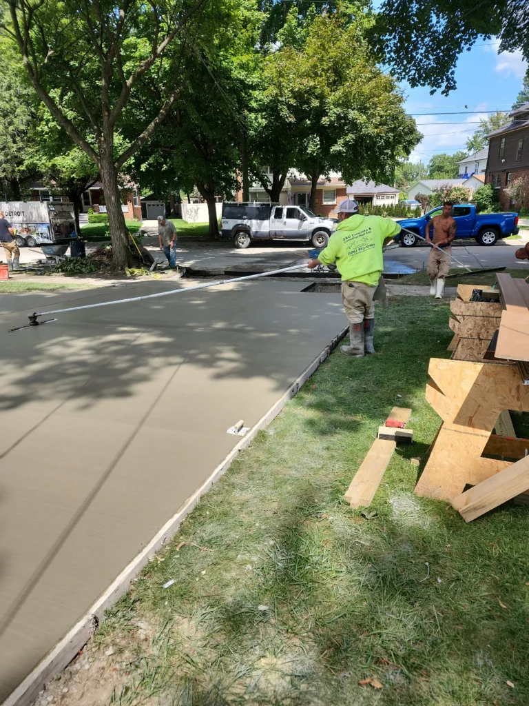 Construction crew lays and smooths a new concrete sidewalk. Worker in yellow uses a bull float on wet concrete.