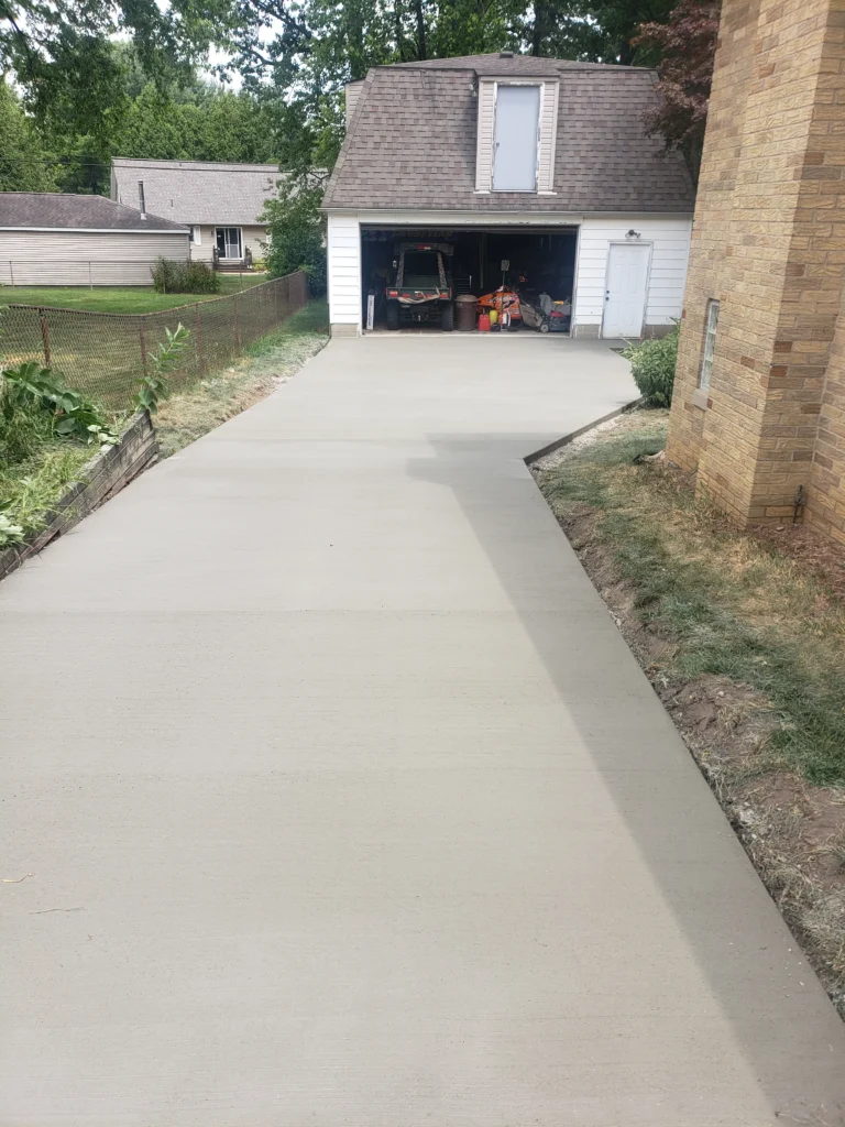 New concrete driveway leading to an open garage showing a utility vehicle and tools. A brick house stands to the right.