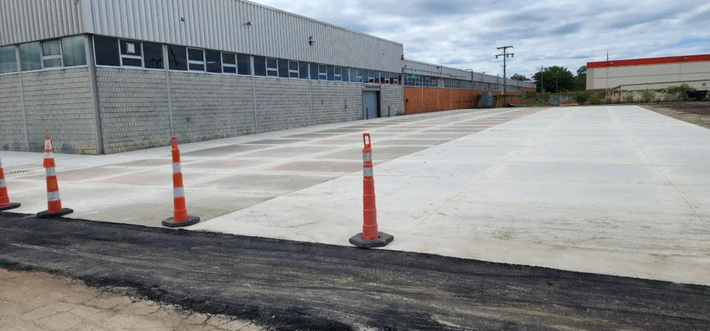 New concrete lot and asphalt strip separated by orange cones, adjacent to an industrial building with windows.