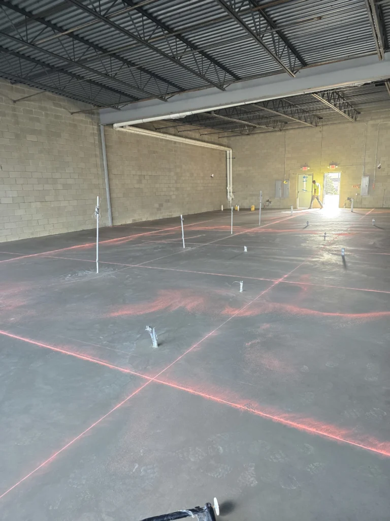 Construction site interior with a concrete floor marked by red laser lines and vertical stakes, and a worker in the distance.