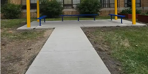 A concrete walkway leads to a patio featuring three blue metal benches and yellow poles, surrounded by grass and bare earth, with a brick building in the background.