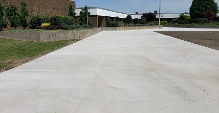Wide concrete path leading towards a building, with a landscaped planter and grass on the left, and a bare earth patch on the right.