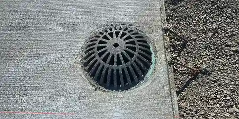 Round storm drain grate embedded in concrete, next to gravel with exposed rebar.