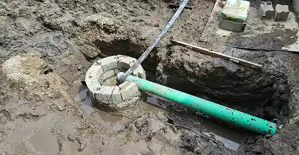 Green drainage pipe installed into a partially built concrete catch basin in a muddy construction trench.